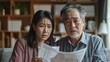 © Pat J. - Asian couple looking anxious and sad, holding bills and discussing financial issues at home