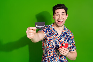 Sticker - Photo of ecstatic guy with stylish haircut dressed print shirt hold smartphone show you credit card isolated on green color background