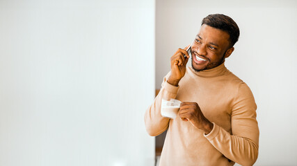 Aufkleber - Cheerful Afro Guy Having Pleasant Phone Conversation Holding Coffee Cup Standing Near Window At Home. Free Space