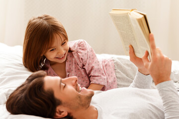 Wall Mural - Young dad reading book to his little daughter, laying on bed together