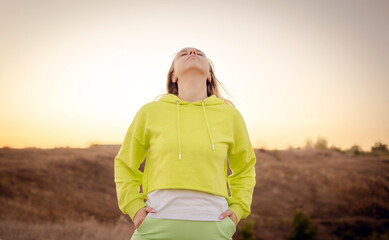 Woman in a bright yellow hoodie stands confidently in a grassy field during sunset, showcasing natural beauty and relaxed style