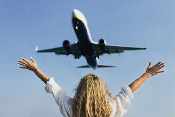 A woman with her arms raised above her head waves at an airplane flying over her