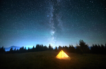 Wall Mural - Glowing yellow touristic tent on grassy hillside under stunning starry sky with Milky Way prominently visible. Silhouette of pine trees and mountains. Serene beauty of clear night in nature.