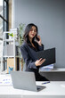 © apichat - A woman is talking on her cell phone while sitting at a desk with two laptops