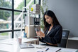 © apichat - A woman is sitting at a desk with a laptop and a cell phone