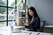 © apichat - A woman is sitting at a desk with a laptop and a cell phone