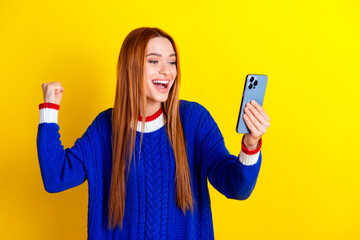 Wall Mural - Photo portrait of lovely young lady hold device winner raise fist dressed stylish blue knitwear garment isolated on yellow color background