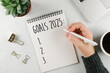 © Anikonaann - Woman's hand writing 2025 Goals in notebook. Laptop, home plant, cup of coffee on light table. Top view, flat lay