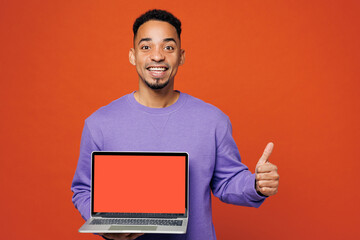 Young IT man of African American ethnicity he wear purple sweatshirt casual clothes hold use work on laptop pc computer show thumb up isolated on plain red orange background studio. Lifestyle concept.