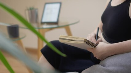 Aufkleber - A woman is sitting at a home office desk, writing in a notepad with a laptop in the background. She is focused and organized in her workspace, creating a productive and professional environment