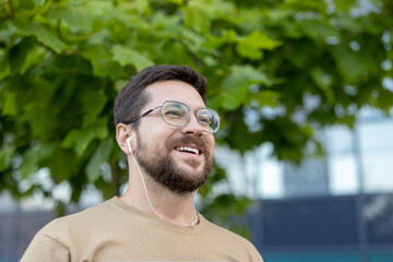 Close-up photo of a young happy man in glasses and headphones who is outside on the street, doing sports and running, smiling and looking ahead