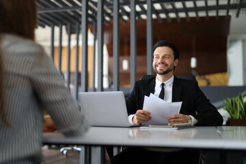 Wall Mural - A man in a suit is sitting at a desk with a woman. He is holding papers and smiling. The woman is sitting across from him, looking at him. The scene appears to be a business meeting or interview