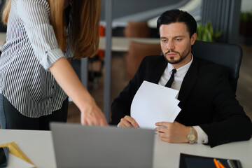 Wall Mural - A man in a suit is sitting at a desk with a woman standing behind him. The man is holding papers and looking at a laptop. The woman is pointing at something on the laptop