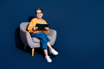 Wall Mural - Full size photo of attractive mature woman sit armchair look empty space read book dressed yellow clothes isolated on dark blue background