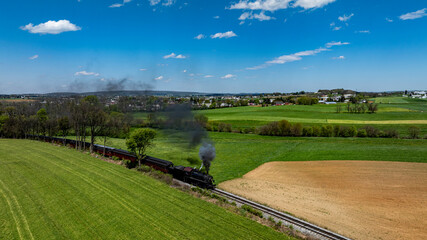 A vintage steam locomotive chugs along the railway, emitting black smoke against a backdrop of vibrant fields and an expansive blue sky, showcasing countryside charm and tranquility.