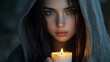 © ifoto - A young woman with long hair in a gray hooded cloak holds a candle, with blue eyes, looking at the camera, against a dark background