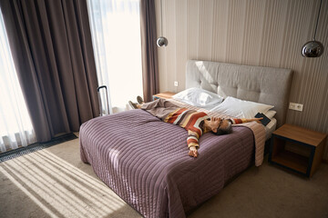 Happy adult woman with suitcase lying the hotel room and falling contentedly on the bed