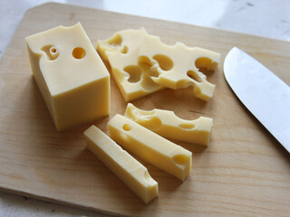 Cheese with holes cut into pieces lies next to a knife on a wooden board on a white background. Cheese making and dairy products. Kitchen, cooking, appetite. Dutch maasdam cheese