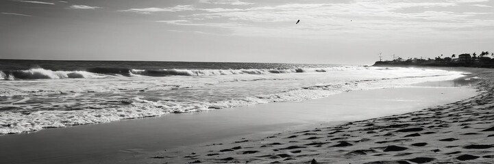 Poster - A black and white photograph beach scene, featuring waves crashing onto the shore, a bird flying overhead, and a distant shoreline with buildings