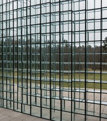 Poster - a modern glass structure composed of numerous rectangular panels arranged in a grid-like pattern, with a blurred background of trees and a grassy area