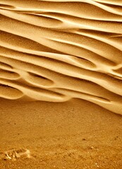 Poster - a close-up view of a sandy beach, with the sand's undulating patterns and ripples created by the wind. The sand is a light brown color, and the background is blurred,