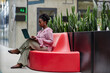 © Seventyfour - Side view of African American businesswoman using laptop to work sitting on red couch in lounge area featuring green live plants in modern office center, copy space