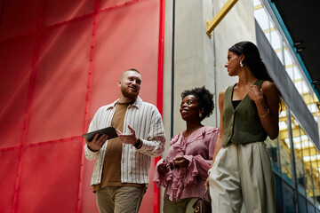 Low angle shot of diverse team of three smiling coworkers discussing business matters while walking together in modern office center, copy space