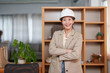 © Ekkasit A Siam - Asian female architect wearing white hard hat smiling while giving thumbs up in office wearing beige blazer bookshelf professional woman in thirties in work environment modern architecture industry