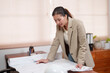 © Ekkasit A Siam - Asian female architect leaning over table, examining blueprints in   office. Wearing beige blazer, concentrating on project details.  office setting is organized with folders and shelves in background