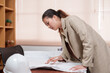 © Ekkasit A Siam - Asian female architect reviewing project plans on table in an office. Wearing beige blazer, focused on work. white safety helmet is on the table, indicating her involvement in construction projects.