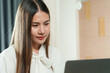 © Ekkasit A Siam - Asian woman working from home sitting at desk wearing white blouse focused on laptop relaxed home office environment modern workspace professional lifestyle indoors.