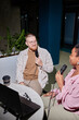 © Seventyfour - Vertical shot of smiling man in casual outfit co hosting podcast with African American woman while creating content using professional microphones at cafe table, camera flash, copy space