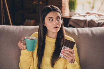 Wall Mural - Photo of charming teen girl sit sofa drink tea eat chocolate look shocked empty space wear yellow clothes modern interior flat indoors