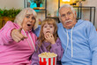 © Andrii Iemelianenko - Shocked Caucasian grandfather, grandmother and granddaughter eating popcorn watching movie on couch at home. Smiling small girl with grandparents enjoying film during weekend in living room apartment