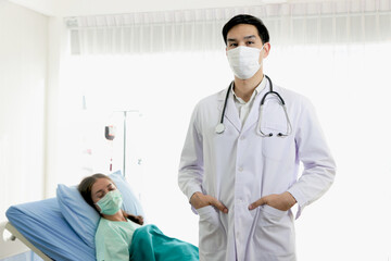 Portrait of young male doctor with stethoscope wearing face mask, standing in white treatment room with the patient lying down on bed as a blurred background. People in the medical field