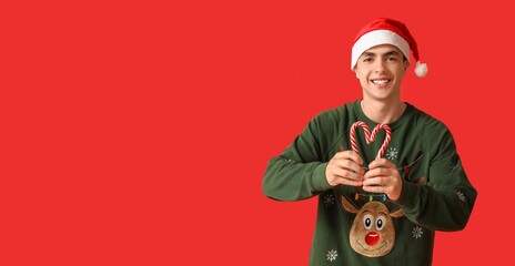 Poster - Happy young man in Christmas sweater and Santa hat holding candy canes on red background with space for text