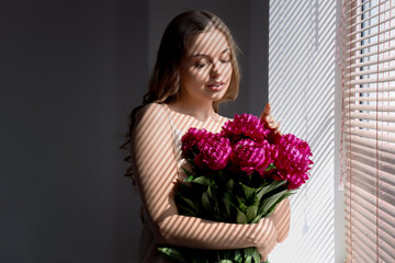 Canvas Print - Beautiful young woman with bouquet of peony flowers near window with blinds