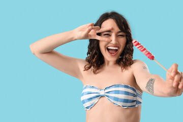 Canvas Print - Young woman in swimsuit with lollipop showing victory gesture on blue background