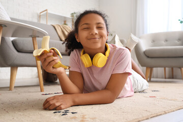 Wall Mural - Little African-American girl eating banana on floor at home