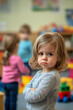© wolfhound911 - A 3-year-old Caucasian girl standing apart in a kindergarten classroom, looking confused and shy, as other children play together in the background with colorful toys.