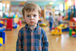 © wolfhound911 - A 3-year-old Caucasian boy standing alone in a colorful kindergarten, looking confused and unsure, surrounded by toys and other children playing in the background.