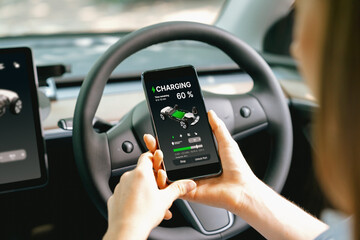 Young woman inside the vehicle, checking EV vehicle's application on battery recharging electricity status display on smartphone screen in modern EV car on her road trip journey. Exalt