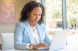 © Tada - woman working on laptop  in her early fourties, wearing business attire with blue blazer and white shirt underneath working on laptop at office table. She has curly hair pulled back into tight curls