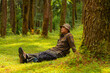 © Yasuspade - An adventurer is sitting on green grass in a lush pine forest area. Asian man in flannel shirt and bucket hat is sitting and resting while enjoying the tranquility of beautiful nature