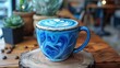 © Suthathip - A vibrant blue spirulina latte served in a ceramic mug, with intricate foam art on top. The mug is set on a wooden table with a modern café background,