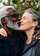 © Daniel - Interracial senior couple, kiss and love in park, garden or nature for kiss or care together. Happy man, woman and partner for retirement, relax and support for marriage and vacation for pensioner