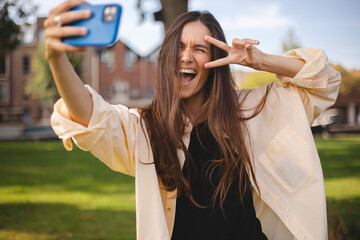 Wall Mural - Image of smiling young brunette hair woman taking selfie photo holding her mobile phone while walking in park. Girl make peace gesture, show two fingers, look happy and smiling, wink or blink.