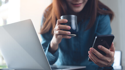 Wall Mural - Young smart business woman taking a coffee break and using mobile phone during working on laptop computer at office. Female freelancer using smartphone and drinking coffee