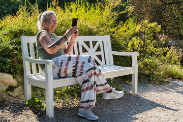 Female tattooed best ager enjoys a sunny autumn day in the park on a bench and takes photos with her smartphone