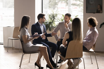 Wall Mural - Serious boss instructing team of employees sitting in circle in office interior. Mentor, coach teaching group of interns. Employees listening to business project leader, writing notes on meeting